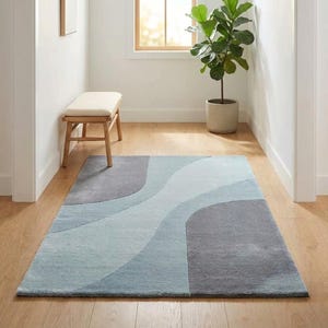 May include: A rectangular area rug featuring a flowing, abstract pattern of large, curved shapes in varying shades of slate gray, light blue, and soft sky blue. The rug has a low-pile texture and is positioned flat on a light-toned wooden floor.