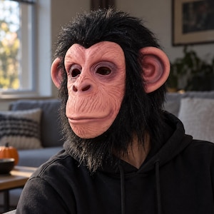 Pode incluir: Uma máscara de chimpanzé realista com um rosto texturizado em rosa claro, órbitas profundas e orelhas proeminentes, rodeada por pelo sintético preto, espesso e desgrenhado, que cobre a cabeça e o pescoço, usada sobre uma camisola preta com capuz.
