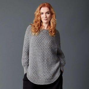 May include: A grey long-sleeved sweater featuring a textured diamond knit pattern, a round neckline, and a curved hemline. The garment has ribbed cuffs and a ribbed collar, providing a structured fit with a consistent heathered grey color throughout.