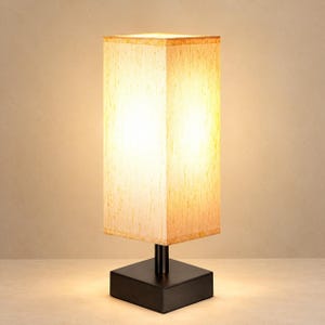 May include: A rectangular table lamp featuring a tall, off-white fabric shade with a subtle textured pattern. The shade is supported by a central black cylindrical post attached to a solid, square black base, emitting a warm, soft glow.