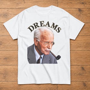 May include: A white short-sleeved t-shirt featuring a graphic print of an older person with white hair, wearing round glasses and a grey suit jacket with a tie, smoking a pipe. The word DREAMS is printed in a black arched font above the portrait.