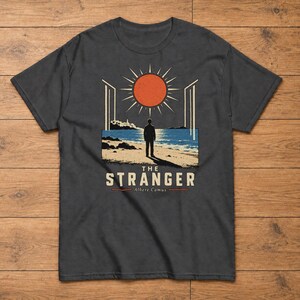Könnte beinhalten: Ein anthrazitfarbenes Kurzarm-T-Shirt mit einem Grafikdruck einer Person, die an einem Strand unter einer großen orangefarbenen Sonne steht. Der Text THE STRANGER und Albert Camus ist unter der Illustration in einer serifenlosen Schrift gedruckt.