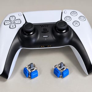 May include: A white and black video game controller with a central PlayStation logo, featuring two analog sticks, a directional pad, and four action buttons, accompanied by two replacement blue and silver joystick sensor modules.