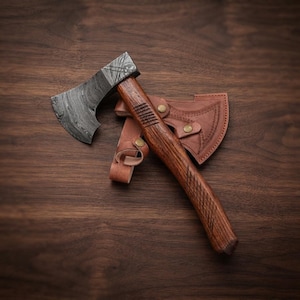 May include: A hand axe with a dark, wavy patterned Damascus steel blade and a curved brown wooden handle featuring textured grip sections. The axe is accompanied by a tan leather sheath with brass snap buttons and decorative stitching.