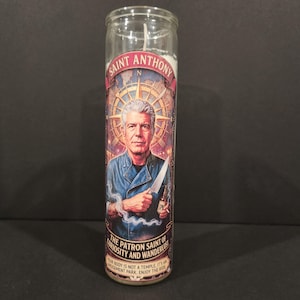 May include: A tall cylindrical glass prayer candle featuring a graphic of Anthony Bourdain in a blue chef coat holding a knife and a bottle. The label reads SAINT ANTHONY, THE PATRON SAINT OF CURIOSITY AND WANDERLUST, and YOUR BODY IS NOT A TEMPLE, IT'S AN AMUSEMENT PARK. ENJOY THE RIDE.