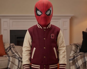 Könnte beinhalten: Eine rote Spider-Man-Maske mit schwarzem Spinnennetz-Muster und weißen Augenlinsen, kombiniert mit einer kastanienbraunen College-Jacke mit cremefarbenen Ärmeln, einem S-Buchstaben-Aufnäher auf der Brust, goldenen Druckknöpfen und gestreiften Rippbündchen.