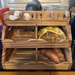 May include: A three-tiered wooden storage rack with a natural wood grain finish. The top shelf holds garlic bulbs, the middle shelf contains a mesh bag of small potatoes, and the bottom shelf holds two sweet potatoes. The text ZelviNest is visible.