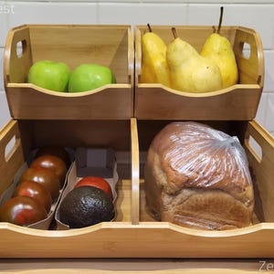 May include: A two-tiered bamboo storage organizer with four open compartments. The top level holds two green apples and three yellow pears. The bottom level contains a row of brown tomatoes, a dark avocado, and a loaf of bread in a plastic bag.