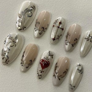 May include: A set of ten almond-shaped press-on nails featuring a glossy off-white and nude base. The designs include metallic silver filigree swirls, a central silver cross with a red gem, and a prominent red heart emblem on one nail.