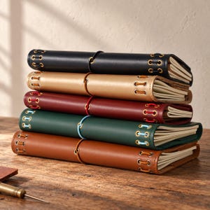 May include: A stack of five leather-bound journals in black, gold, burgundy, dark green, and brown. Each journal features metal eyelets along the spine with colored lacing and a central elastic band closure holding the pages securely in place.