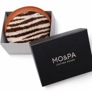 May include: A round coin purse with a brown and white zebra-striped hair-on-hide pattern and a tan leather zipper border, nestled inside a light gray box next to a black lid featuring the white text MO&PA LEATHER GOODS printed on the top surface.