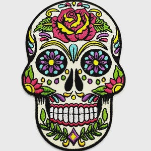 May include: An embroidered patch shaped like a human skull with a cream-colored base. The design features a large pink rose on the forehead, purple and blue floral eye sockets, green leaves, and yellow accents, all outlined in thick black thread.
