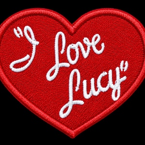 May include: A heart-shaped embroidered patch with a bright red background and a thick red stitched border. The white embroidered text reads "I Love Lucy" in a cursive script font, centered across the middle of the heart-shaped fabric surface.