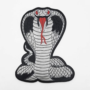 May include: An embroidered patch featuring a cobra snake with a flared hood. The design uses silver-grey thread for the scales, black thread for the hood and outline, and vibrant red thread for the snake's eyes and its forked tongue.