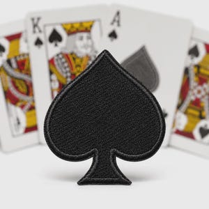 May include: A solid black embroidered patch in the shape of a spade suit symbol from a deck of cards. The patch features a dense, textured thread construction with a clean, stitched border, positioned in front of blurred playing cards.