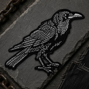 May include: An embroidered patch featuring a black raven with detailed white stitching that highlights the bird's feathers, beak, and eye. The raven is depicted in profile, facing right, with its wings folded against its body and talons visible.