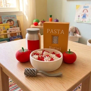 Può includere: Un set di giocattoli in legno composto da una ciotola bianca riempita con forme di pasta rosse e bianche, una scatola gialla con l'etichetta PASTA Rotini, un barattolo argentato con etichetta rossa, due pomodori rossi e un mestolo per pasta grigio.