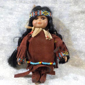 May include: A vintage Native American doll with black hair and a brown suede outfit. The doll has a beaded headband and a necklace. The doll's outfit has fringe on the sleeves and legs. The doll is standing on a white surface with a floral pattern.
