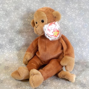 May include: A plush monkey toy in shades of brown and tan, with a tag attached. The monkey is sitting on a patterned surface. The toy has a soft, cuddly appearance, with detailed features.