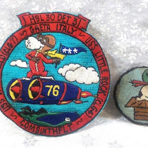 May include: Two embroidered patches, one blue with a cartoon dog flying a plane and the text "SPRINGFIELD (CLG7) HSL 30 DET BI GAETA ITALY USS LITTLE ROCK (CLG4) LA COMSIXTHELT 2" and the other a brown and green patch with a cartoon dog wearing a pilot's helmet.