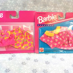 May include: Two Barbie Fashion Favorites outfits in their original packaging. One outfit is a pink top and skirt with a yellow floral pattern. The other outfit is a pink top and skirt with a yellow and pink floral pattern.