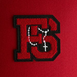 May include: A red and black embroidered patch in the shape of the letter B. The patch features a textured red center with a black border. A white rosary with a small cross is stitched onto the center of the letter, overlaying the red fabric.