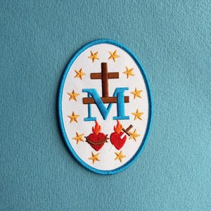 以下が含まれることがあります： 白い背景と青い縁取りの楕円形の刺繍ワッペン。茶色の十字架、大きな青い文字のM、炎のある2つの赤いハートが描かれています。中央のシンボルの周りには12個の黄色い星が円状に配置されています。