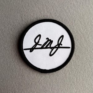 May include: A circular embroidered patch featuring a white fabric background with a black stitched border. The centre displays a black cursive signature or stylised lettering that reads JmJ, with a horizontal line extending across the middle.