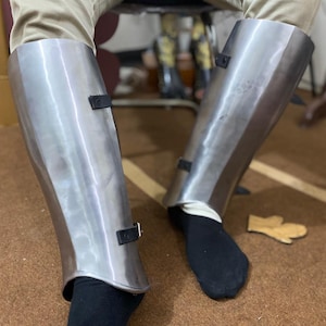 Puede incluir: Un par de grebas de acero pulido diseñadas para proteger la parte inferior de las piernas. Cada pieza de armadura presenta una superficie metálica lisa y curva, asegurada con dos correas de cuero negro con hebillas de metal plateado.