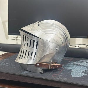 Peut inclure: Un casque de chevalier en métal argenté poli doté d'une visière articulée avec des fentes de ventilation verticales, des motifs gravés complexes et une mentonnière en cuir marron avec une boucle en métal fixée sur le côté.