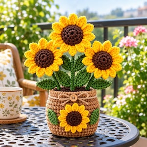 May include: A crochet sunflower arrangement featuring three bright yellow flowers with dark brown centres and green leaves, set in a textured brown pot decorated with a small sunflower applique, a jute twine bow, and a single wooden button.