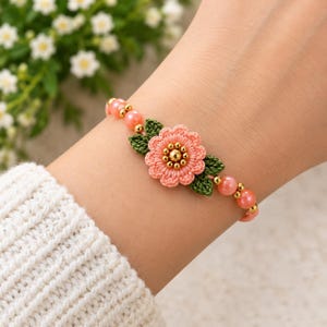 Puede incluir: Una pulsera con una flor bordada central de color melocotón con un centro de cuentas doradas, flanqueada por dos hojas bordadas de color verde oscuro. La banda consta de cuentas redondas de color melocotón y pequeñas cuentas metálicas doradas.