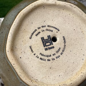 Puede incluir: Vista inferior de una maceta de cer&aacute;mica con un exterior verde terroso moteado y un interior beige. La base presenta el texto "Handwash Do Not Microwave Made in China" y "UO Home".