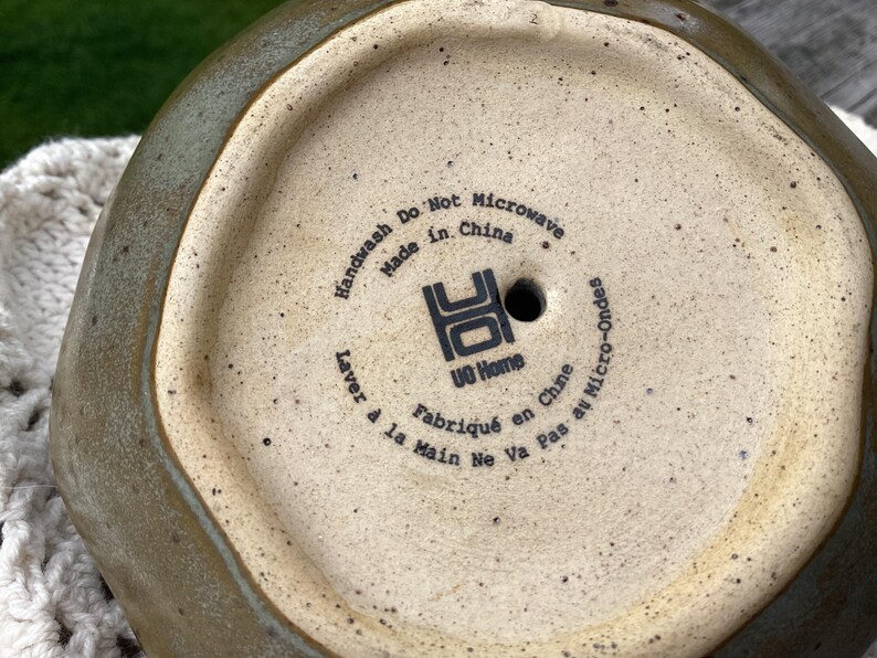 Puede incluir: Vista inferior de una maceta de cer&aacute;mica con un exterior verde terroso moteado y un interior beige. La base presenta el texto "Handwash Do Not Microwave Made in China" y "UO Home".