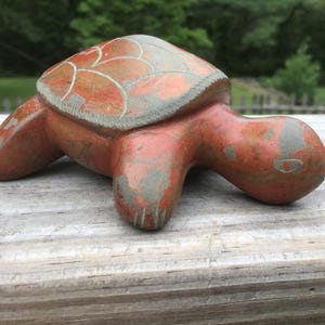 May include: A terracotta-colored turtle figurine with a weathered, matte finish. The shell features etched, light-colored line patterns forming a geometric design. The turtle has a smooth, rounded head and four legs, with visible signs of surface wear.