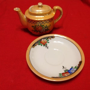 May include: A miniature orange and gold teapot with a matching saucer. The teapot has a lid and handle, and both pieces are decorated with floral patterns in red, blue, and green. The saucer has a white center.