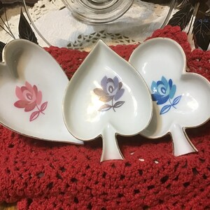 May include: Three small ceramic trinket dishes shaped like hearts and a spade. The dishes are white with gold trim and feature a floral design in pink, blue, and purple.