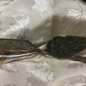 May include: Two antique silver serving utensils, a cake server and a pie server, rest on a white tablecloth. Both utensils have ornate floral patterns. The cake server has a rectangular blade with a serrated edge, while the pie server has a rounded, leaf-shaped blade.