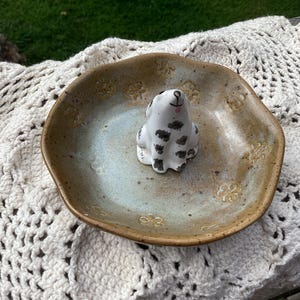 Puede incluir: Un plato de cer&aacute;mica con un centro azul claro y un borde marr&oacute;n, con una peque&ntilde;a figura de perro blanco con manchas negras. El plato tiene un patr&oacute;n floral y est&aacute; sobre una superficie blanca de ganchillo.