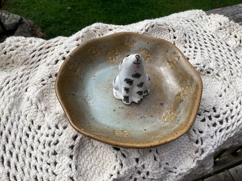 Puede incluir: Un plato de cer&aacute;mica con un centro azul claro y un borde marr&oacute;n, con una peque&ntilde;a figura de perro blanco con manchas negras. El plato tiene un patr&oacute;n floral y est&aacute; sobre una superficie blanca de ganchillo.