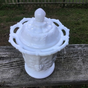 May include: White milk glass candy dish with a lid. The dish is decorated with a grapevine pattern and has a scalloped edge.