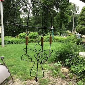 May include: A black metal angel wind chime with a heart design hangs from a metal frame in a garden setting. The chime has wooden beads and is surrounded by green foliage.