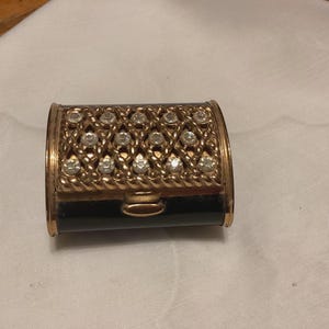 May include: A small, rectangular, vintage compact with a gold-tone metal frame. The top is adorned with a lattice pattern of clear rhinestones and a rope-like border. The base is black.