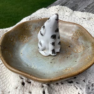 Puede incluir: Un plato de cer&aacute;mica con un esmalte moteado, que contiene una peque&ntilde;a figura blanca con manchas negras. El plato tiene un centro azul claro y bordes marrones, con dise&ntilde;os florales. La figura es un peque&ntilde;o animal sentado.