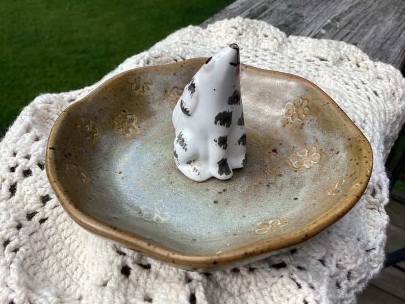 Puede incluir: Un plato de cer&aacute;mica con un esmalte moteado, que contiene una peque&ntilde;a figura blanca con manchas negras. El plato tiene un centro azul claro y bordes marrones, con dise&ntilde;os florales. La figura es un peque&ntilde;o animal sentado.