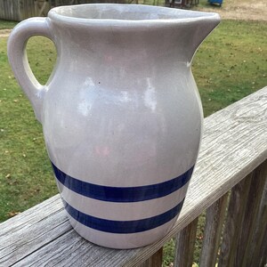 May include: A ceramic pitcher with a light gray glaze and two horizontal blue stripes. The pitcher has a handle and a spout for pouring. It is sitting on a wooden railing.