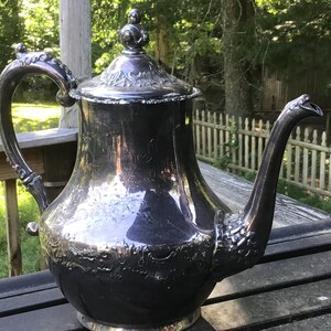May include: A silver teapot with an ornate design and a decorative knob on the lid. The teapot is sitting on a wooden surface.