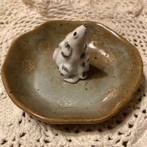 Puede incluir: Un plato de cer&aacute;mica con un esmalte azul claro y marr&oacute;n, con una peque&ntilde;a figura blanca con manchas negras. El plato tiene una forma ligeramente irregular y grabados florales. La figura es un animal peque&ntilde;o.