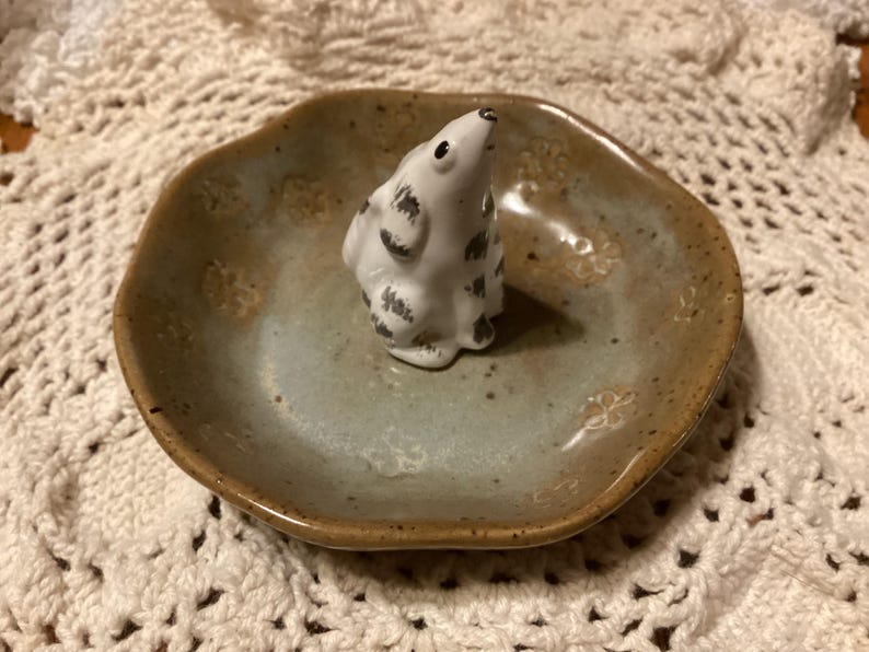 Puede incluir: Un plato de cer&aacute;mica con un esmalte azul claro y marr&oacute;n, con una peque&ntilde;a figura blanca con manchas negras. El plato tiene una forma ligeramente irregular y grabados florales. La figura es un animal peque&ntilde;o.