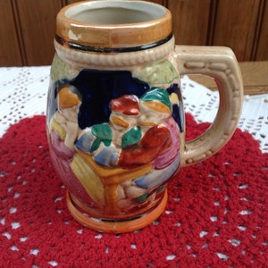 May include: A ceramic beer stein with a colorful scene of three people sitting at a table. The stein has a brown handle and a brown rim.
